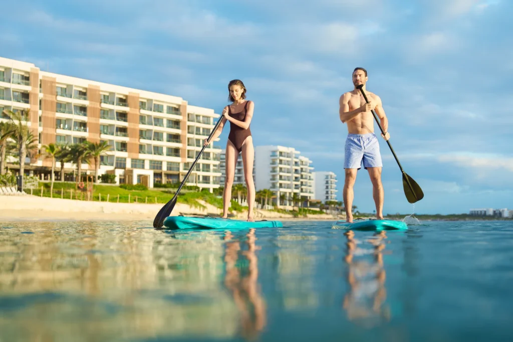 A couple paddle surfing on the beach at Waldorf Astoria Cancun, with the hotel in the background and the ocean surrounding them.