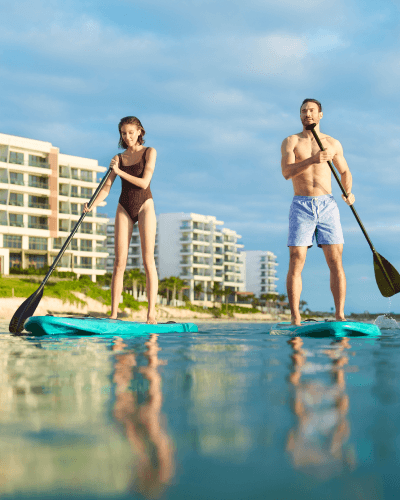 Una pareja practicando paddle surf en la playa del Waldorf Astoria Cancún, con el hotel al fondo y el océano rodeándoles.