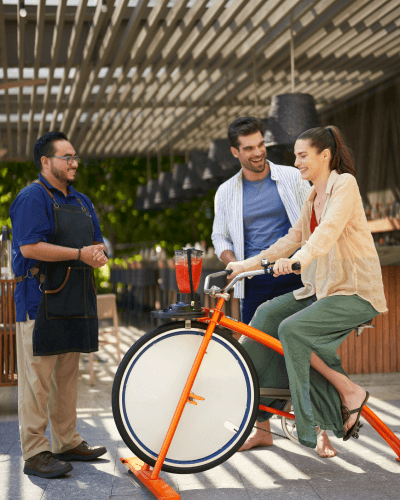 Una pareja se divierte preparando su propia bebida en una batidora, pedaleando para impulsarla mientras disfrutan juntos del proceso.