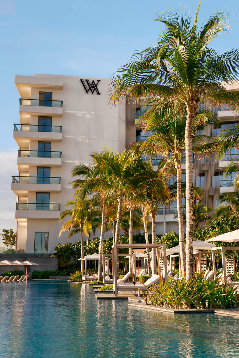 La fachada del Waldorf Astoria Cancun Resort vista desde la piscina, rodeada de palmeras y camas balinesas, creando un ambiente lujoso y tranquilo.