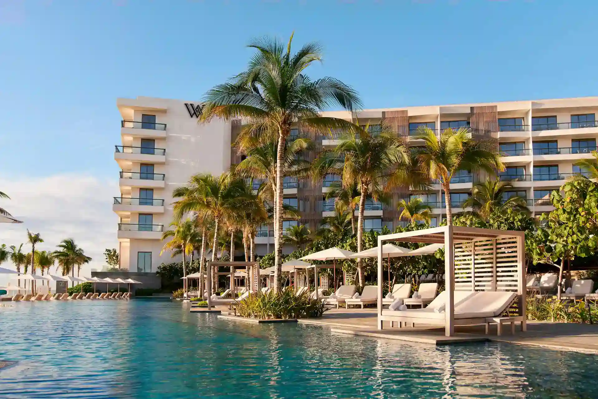 Piscina rodeada de palmeras con tumbonas y cabañas frente al hotel.