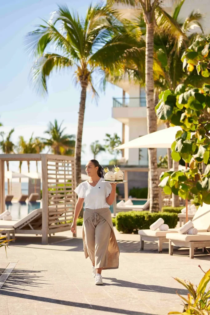 Waldorf Astoria Cancun waitress gracefully serving cocktails by the pool, surrounded by palm trees and sun loungers.
