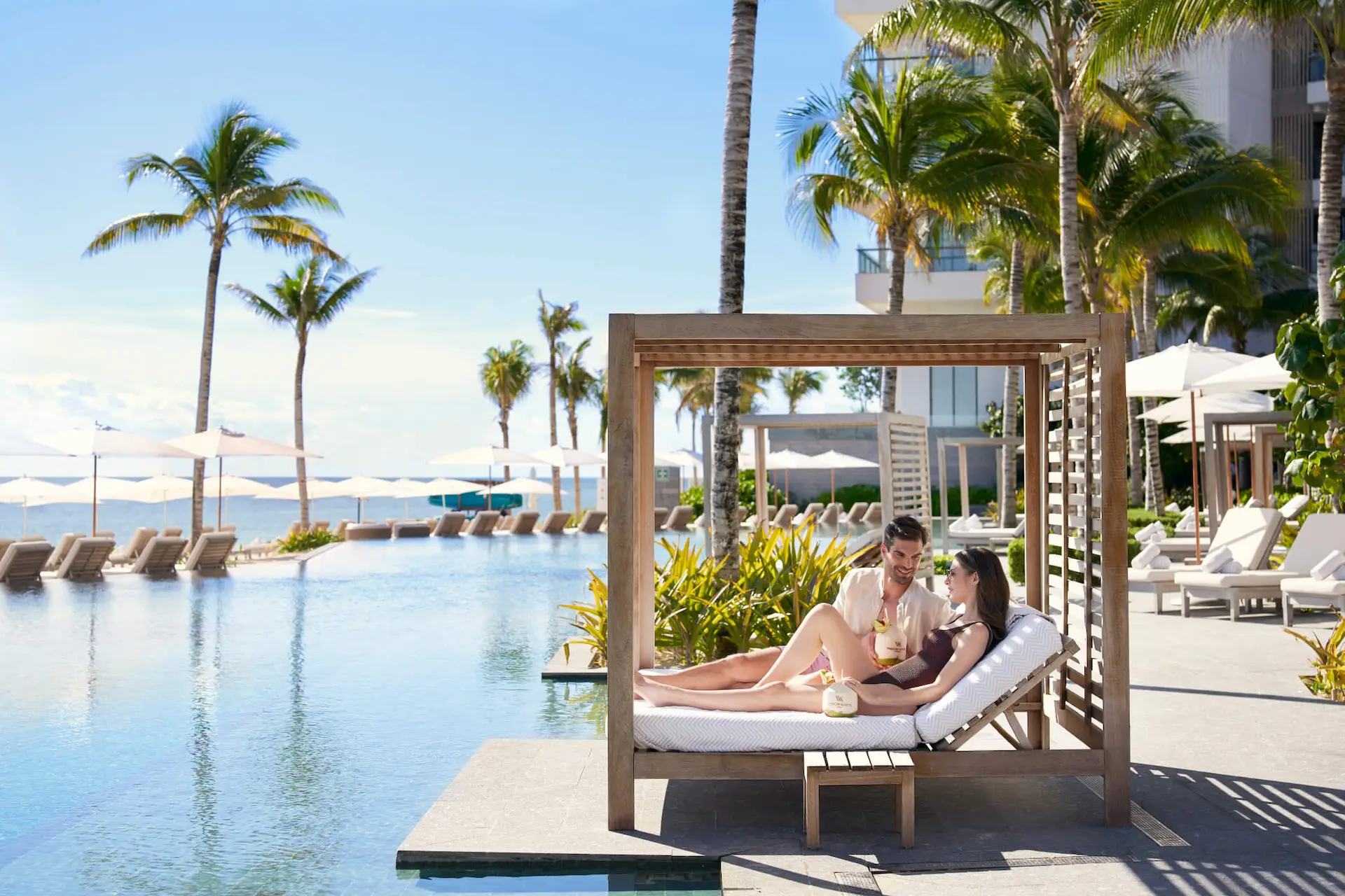 A couple relaxing on a Balinese bed at the edge of the infinity pool, enjoying exquisite cocktails on a splendid day at Waldorf Astoria Cancun.