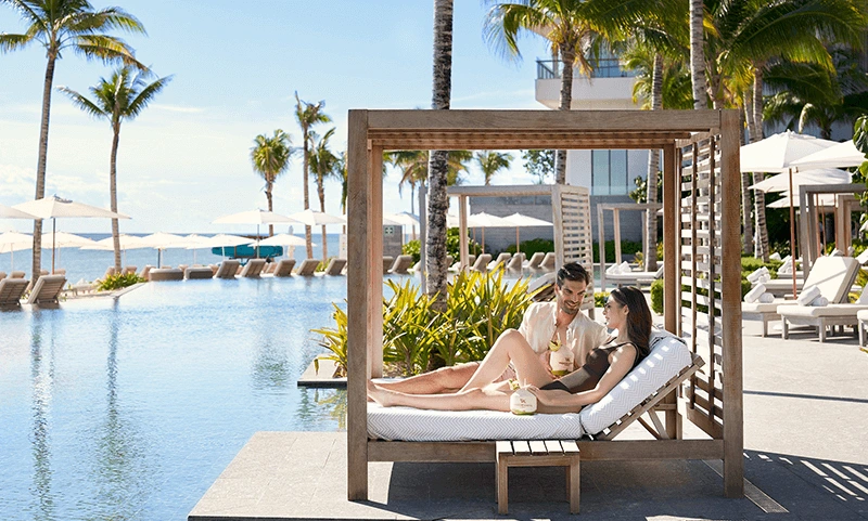 A couple relaxing on a Balinese bed at the edge of the infinity pool, enjoying exquisite cocktails on a splendid day at Waldorf Astoria Cancun.